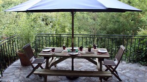 Une table de patio surmontée d’un grand parasol, entourée de chaises et d’un banc, installée sur une terrasse en pierre avec une rambarde en métal.