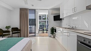 A modern bedroom with a bed, a small sitting area with chairs, a balcony with a table and chairs, and a kitchenette with a TV and appliances.