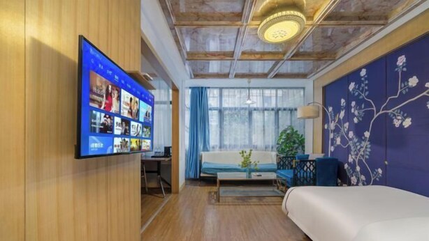 Una camera d'albergo moderna con una grande televisione a schermo piatto fissata al muro, pavimento in legno, un letto, un divano e una parete decorata con motivi floreali.