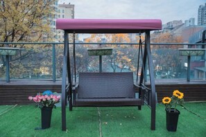 A swing with a canopy, a potted plant with pink flowers, and another with yellow flowers on a balcony with a city view. - Hangzhou Yue Boutique Hotel (Longhu Zijing Tianjie Sandun Subway Station) (Hangzhou)