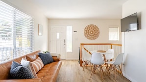 Una sala de estar con un sofá de cuero marrón, sillas blancas, una pequeña mesa de comedor y un televisor montado en la pared.