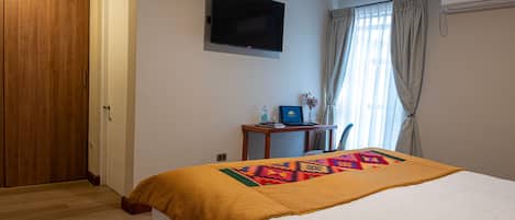 Habitación de hotel con cama, televisor montado en la pared, escritorio con computadora y ventana con cortinas.