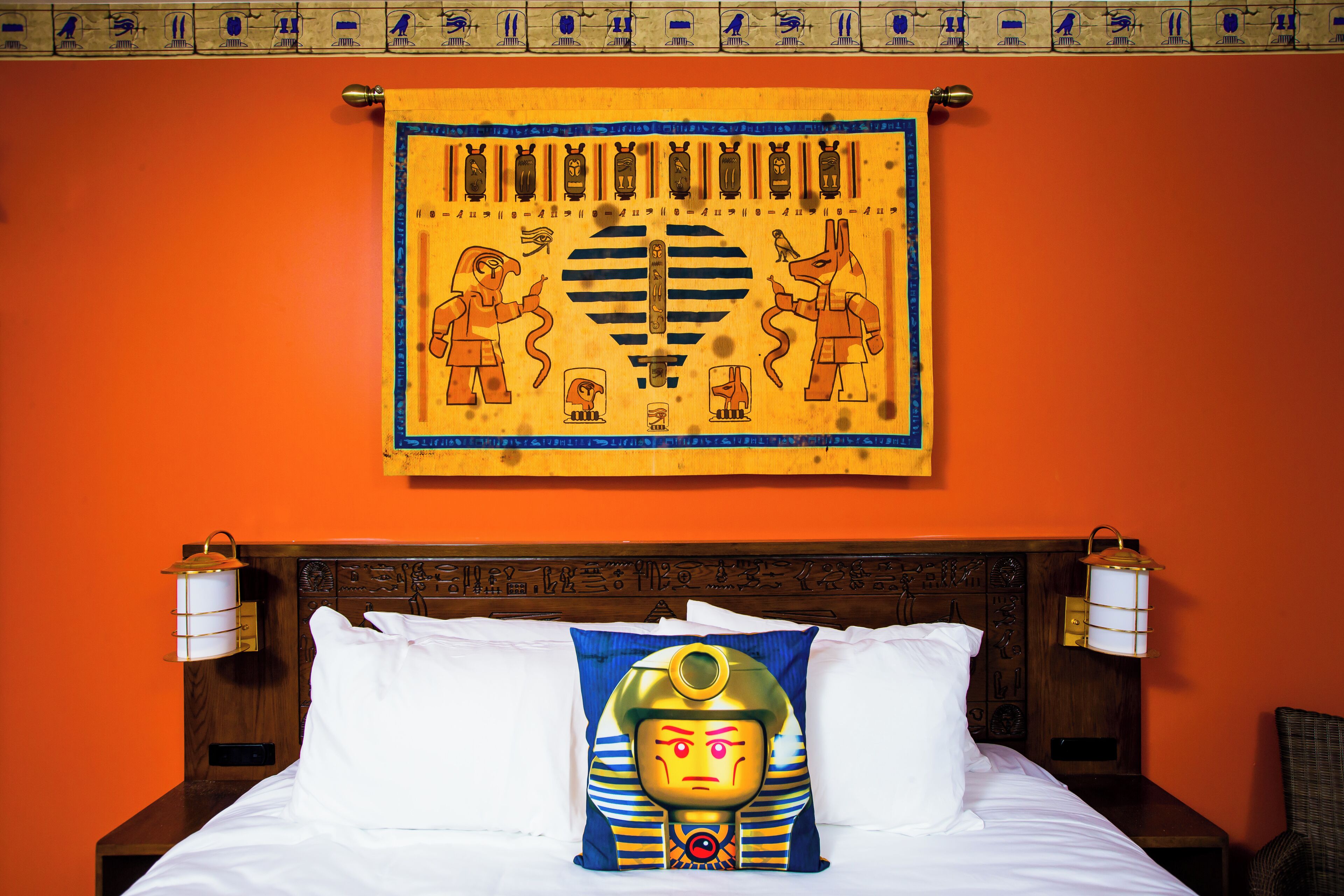 Una cama con sábanas blancas, un almohadón con diseño de temática egipcia y un tapiz mural con jeroglíficos.