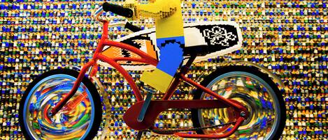 A LEGO figure on a bicycle with a pixelated design on the seat, set against a background of numerous small LEGO figures.