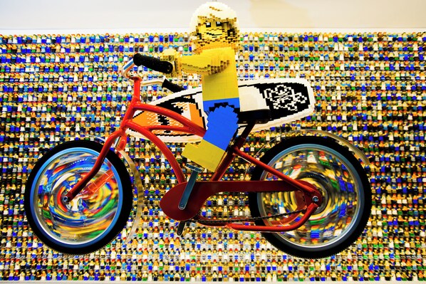 A LEGO figure on a bicycle with a pixelated design on the seat, set against a background of numerous small LEGO figures. - Legoland Florida Hotel (Winter Haven)