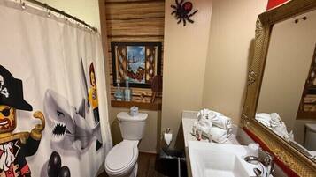Un baño con cortina de ducha decorada con piratas y tiburones, un inodoro, un lavamanos con toallas y un espejo grande.