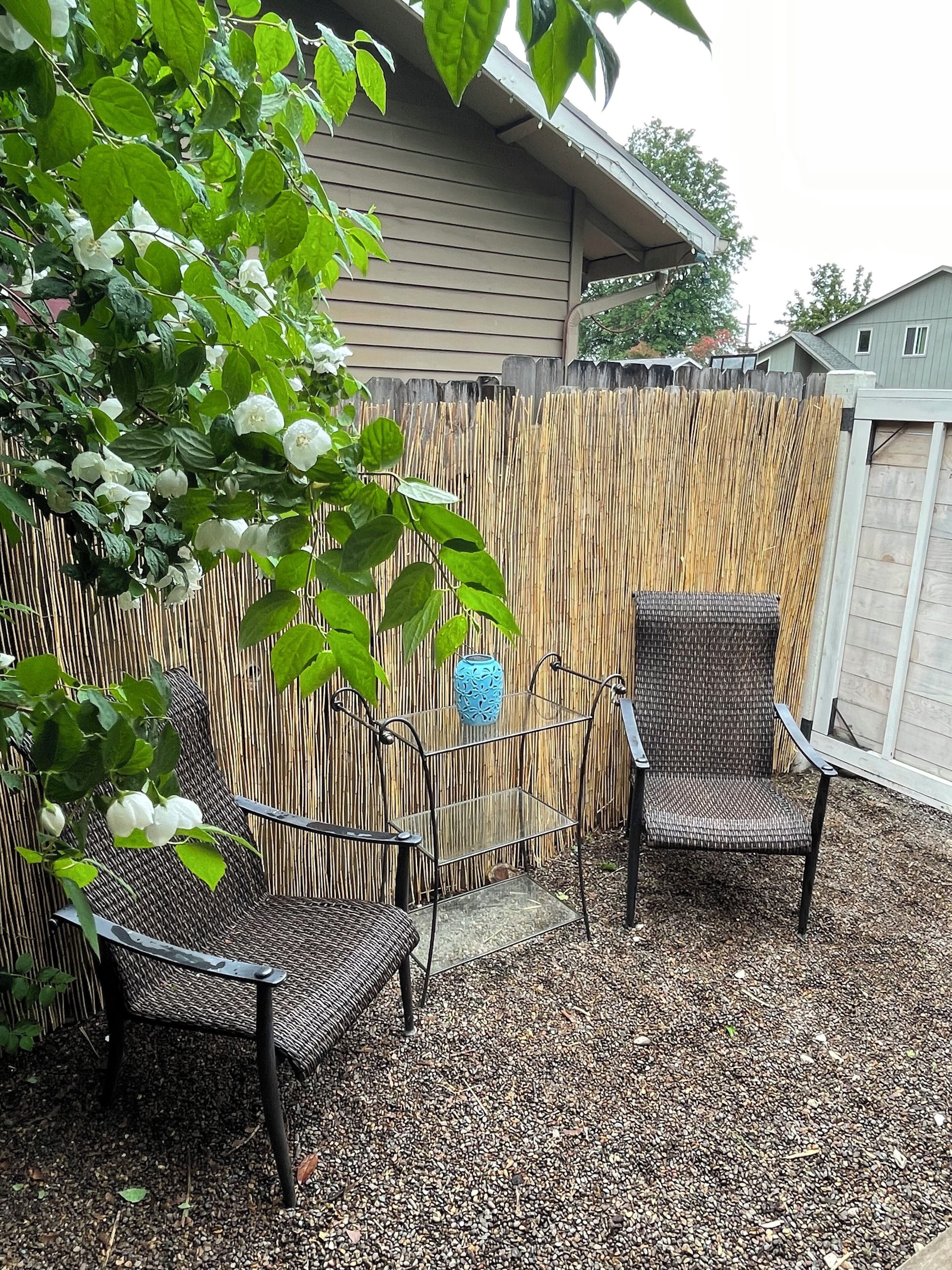 A patio area with two wicker chairs and a small table, surrounded by a bamboo fence and a house in the background.