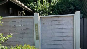 A wooden gate with a 'WELCOME' sign, surrounded by greenery and a building in the background.