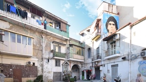 Una calle estrecha flanqueada por edificios, con balcones llenos de ropa, una pequeña tienda y un mural en uno de los edificios.