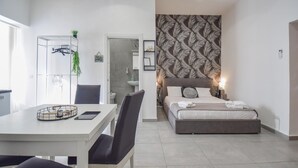 Un dormitorio moderno con cama, mesita de noche y escritorio con silla. Se observa un baño al fondo.