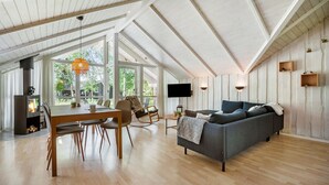 A spacious living area with a high ceiling, wooden beams, a grey sofa, a dining table with chairs, and a TV mounted on the wall.