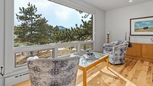 Amplia sala de estar con ventanas amplias que ofrecen vistas a un bosque y montañas. Se observa un sillón con estampado, una mesita con un libro y auriculares, y un aparador de madera.