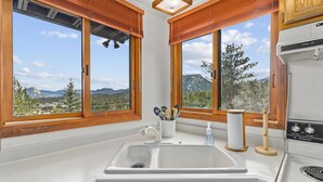 A kitchen with a white sink, wooden cabinets, and a stove. There are two windows with wooden blinds, offering a view of mountains and trees. - Elkhorn West -Amazing views! Close to town! (Estes Park)