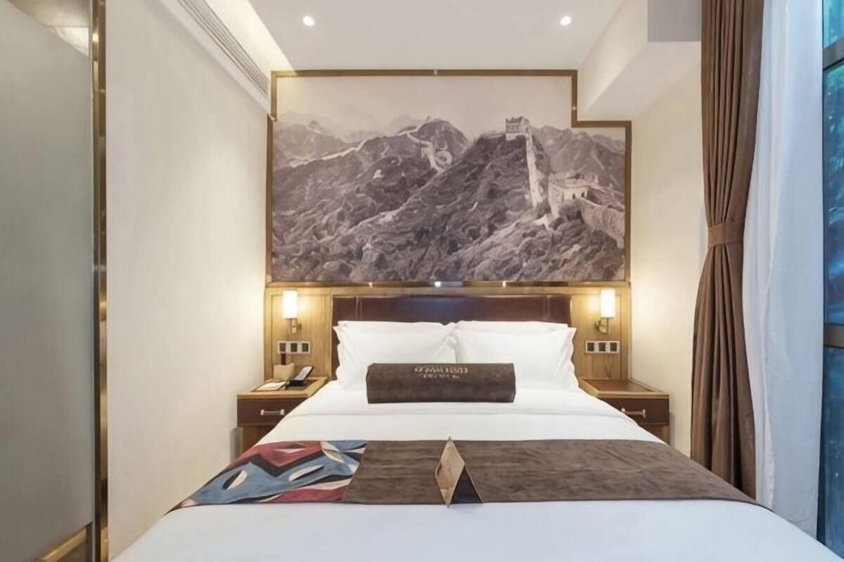 A hotel room with a large bed, two bedside tables, a nightstand, a wall-mounted TV, and a large framed picture of a mountainous landscape.