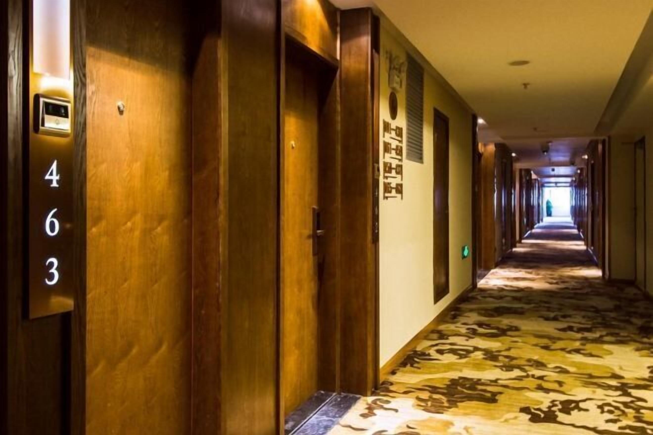 Un pasillo de hotel con puertas de madera, una alfombra estampada y un largo corredor que conduce a un final luminoso.