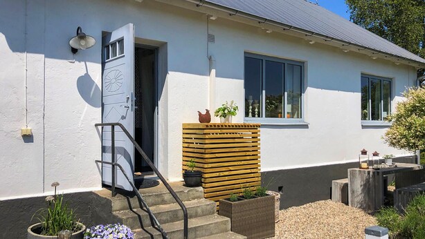 A house with a white exterior, a wooden staircase leading to a door, and a small garden area with potted plants and a wooden fence.