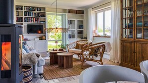 A cozy living room with a fireplace, bookshelves, wicker furniture, and a view of greenery outside.