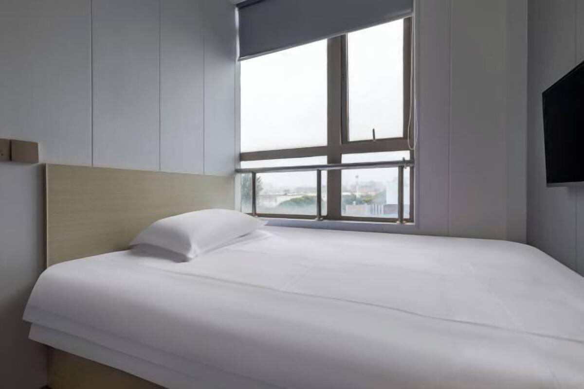 A hotel room with a large window, a bed with white bedding, and a mounted TV on the wall.