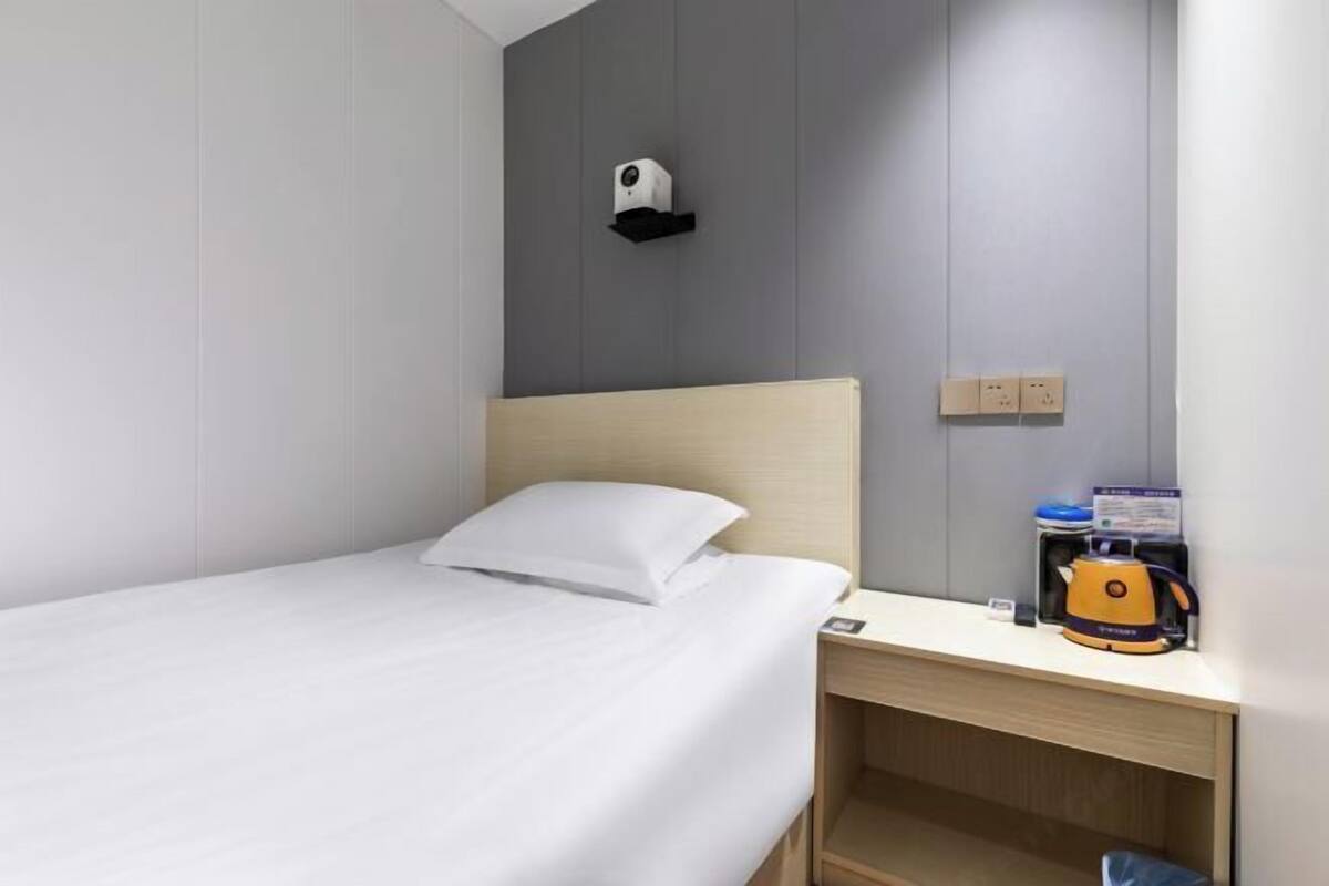 A hotel room with a bed, a nightstand, a kettle, and a security camera.