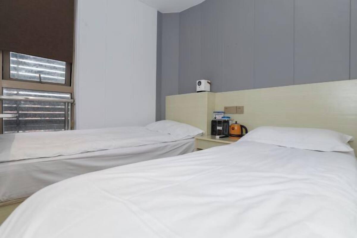 A hotel room with two single beds, a bedside table with a coffee maker, a small cabinet, and a window with blinds.