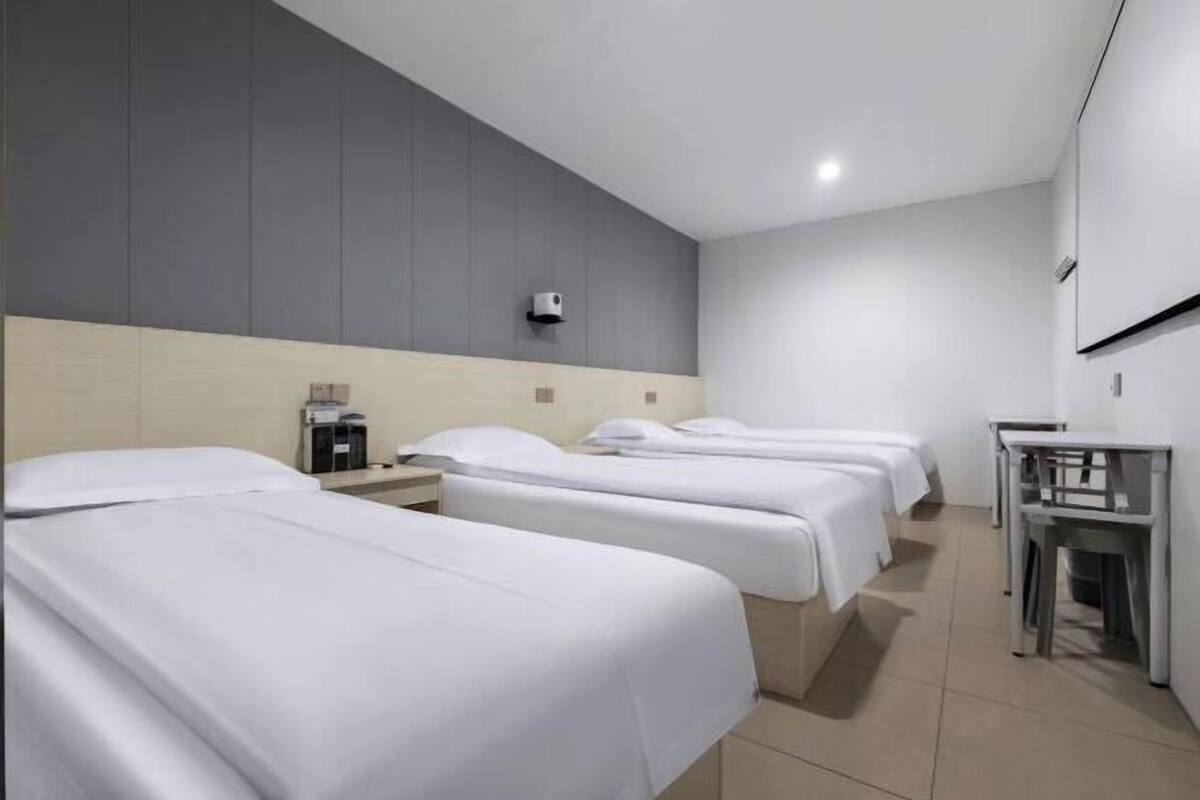 A hotel room with three single beds, a small table, a chair, and a wall-mounted air conditioning unit.