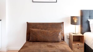 A modern bedroom with a brown leather sofa, a wooden nightstand with a lamp, and a framed abstract artwork on the wall. - VIVA Bartolomeu Beach Flats 3E (Porto)