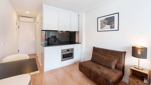 A compact living space with a kitchenette, a brown sofa, a small table, and a lamp. - VIVA Bartolomeu Beach Flats 3E (Porto)
