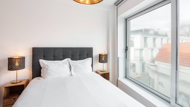 A hotel room with a large bed, two bedside tables, a modern chandelier, and a large window offering a view of buildings. - VIVA Bartolomeu Beach Flats 3E (Porto)