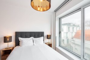 A hotel room with a large bed, two bedside tables, a modern chandelier, and a large window offering a view of buildings.