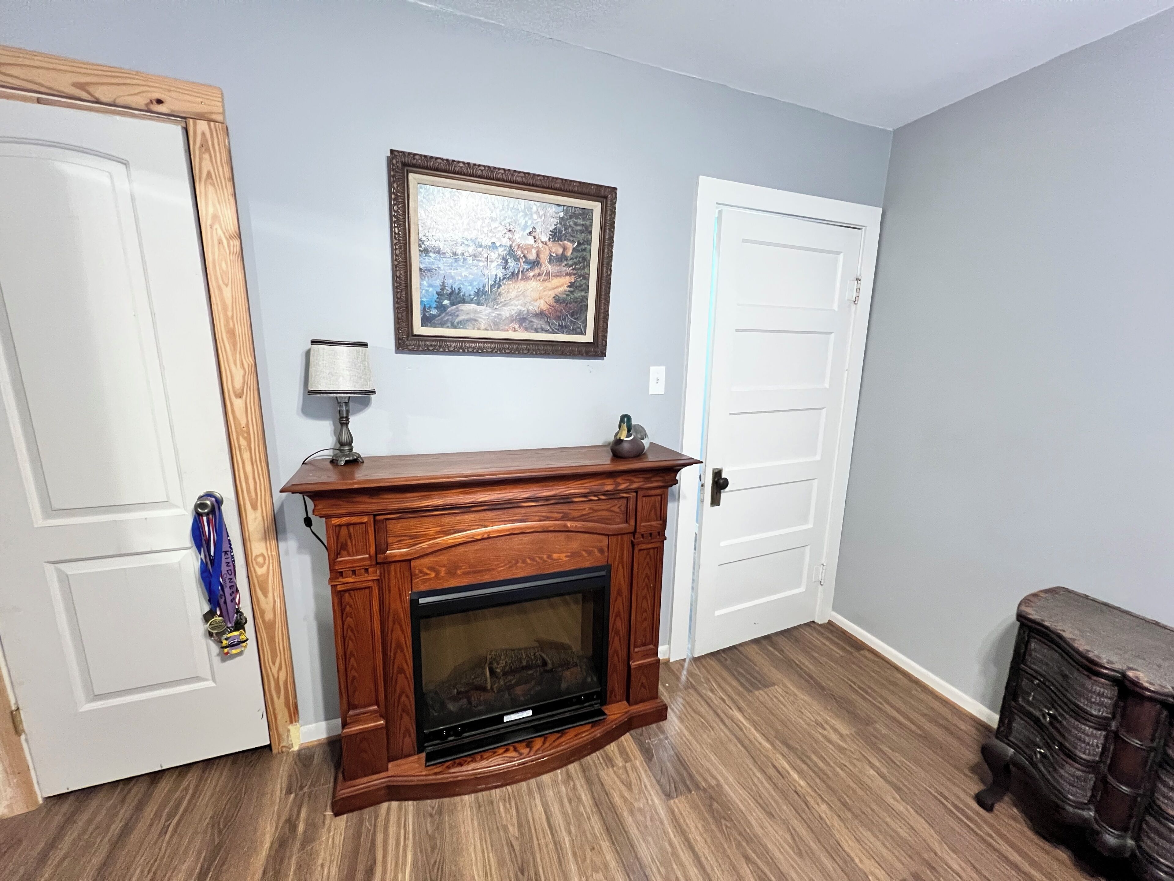 A room with a wooden fireplace, a framed painting above it, and a door on the right.
