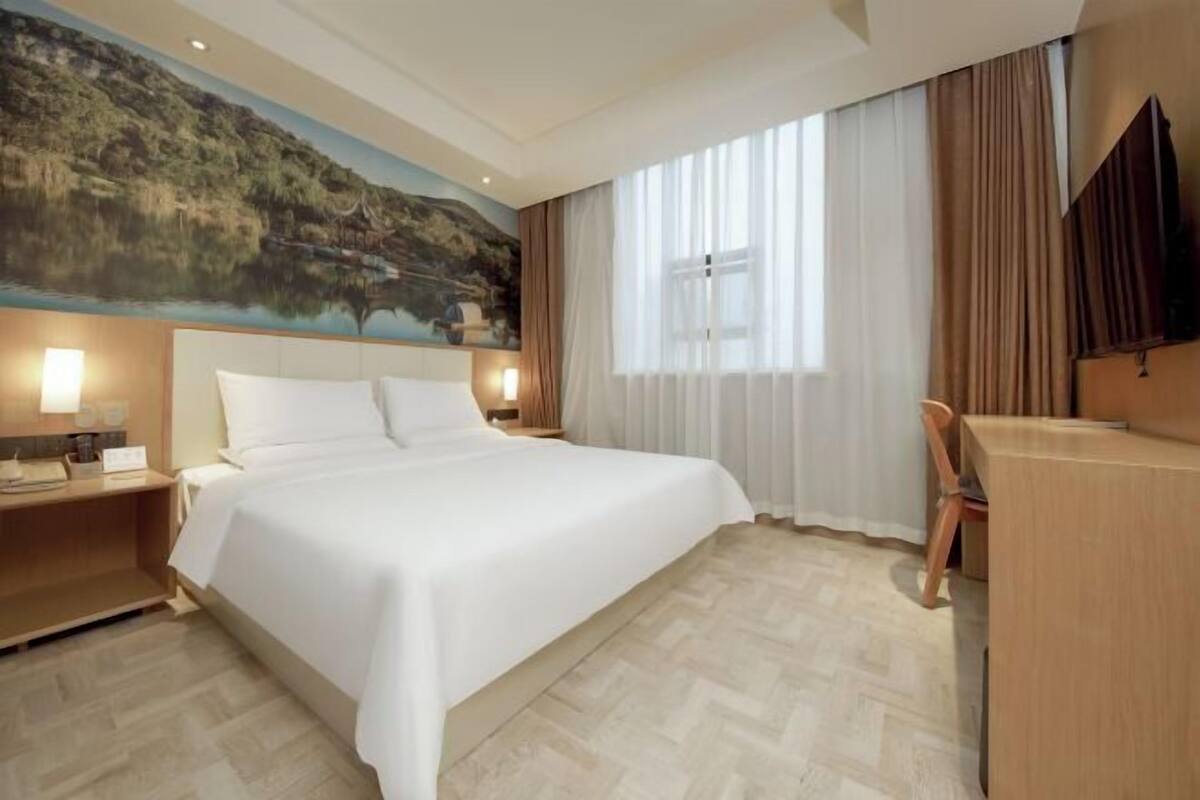 A hotel room with a large bed, a desk, a chair, a painting on the wall, and a window with curtains.