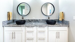 Un baño con dos lavamanos, cada uno con un espejo redondo y un lavabo tipo cuenco negro. Hay armarios blancos con tiradores plateados y una encimera de mármol.