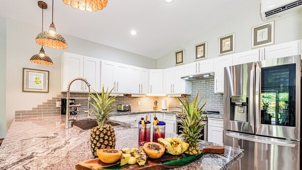 Una cocina con armarios blancos, encimera de granito, refrigerador de acero inoxidable y una exhibición de frutas.