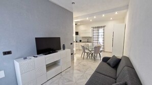 Una sala moderna con un televisor de pantalla plana sobre un mueble blanco, un sofá gris y una zona de comedor con una mesa de vidrio y sillas.