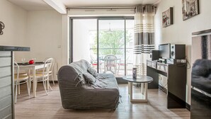 A modern living room with a grey sofa, a dining table with chairs, a TV, and a balcony with a view of greenery.
