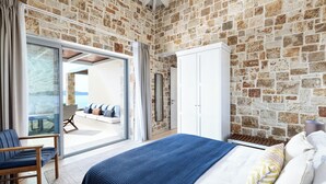 Un dormitorio con pared de piedra, una cama con manta azul, un armario blanco y un ventanal de vidrio que da a un balcón.