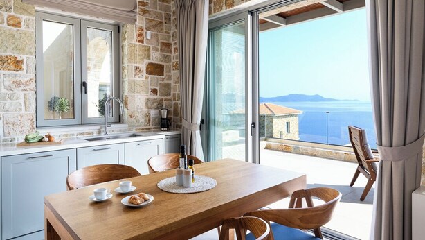 Una cocina con pared de piedra, un ventanal con vistas al mar y una zona de comedor con mesa y sillas de madera.