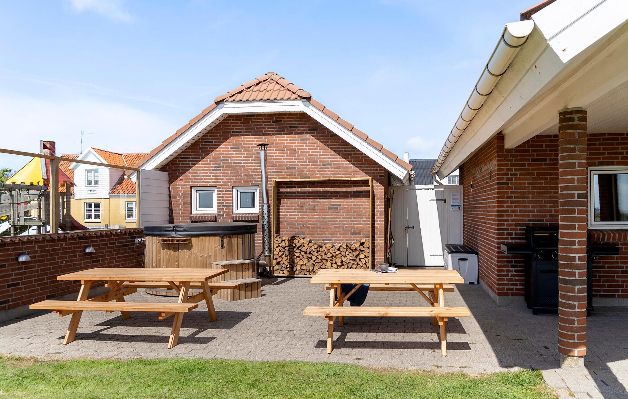 A brick building with a pitched roof, a wooden picnic table, a grill, and a stack of firewood.