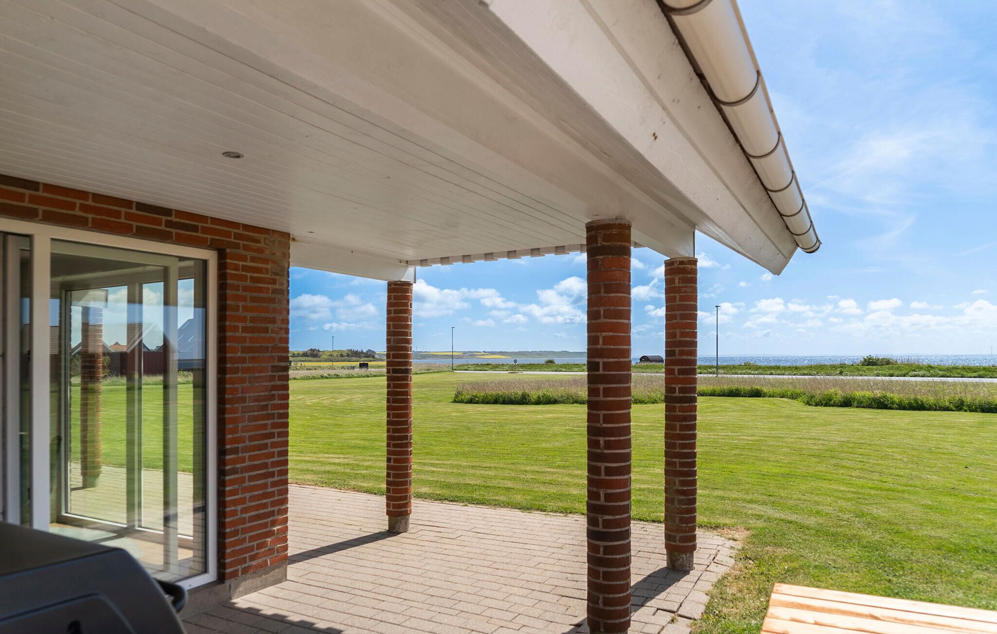 Eine überdachte Terrasse mit Ziegelstützen, einer Glastür und Blick auf ein Wiesfeld und Wasser.
