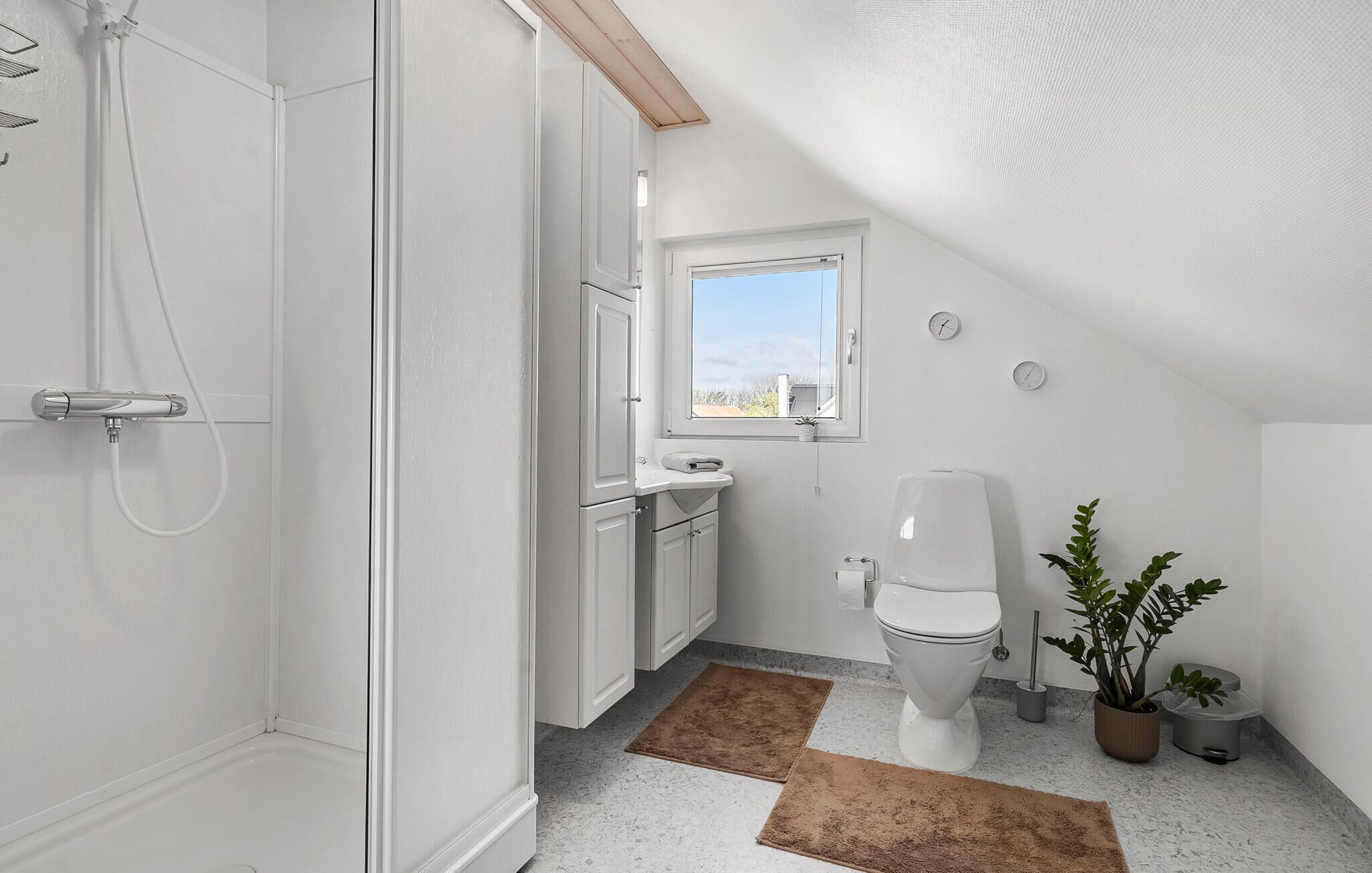 A small, white bathroom with a shower, toilet, and sink. There is a window providing natural light and a plant on the floor.