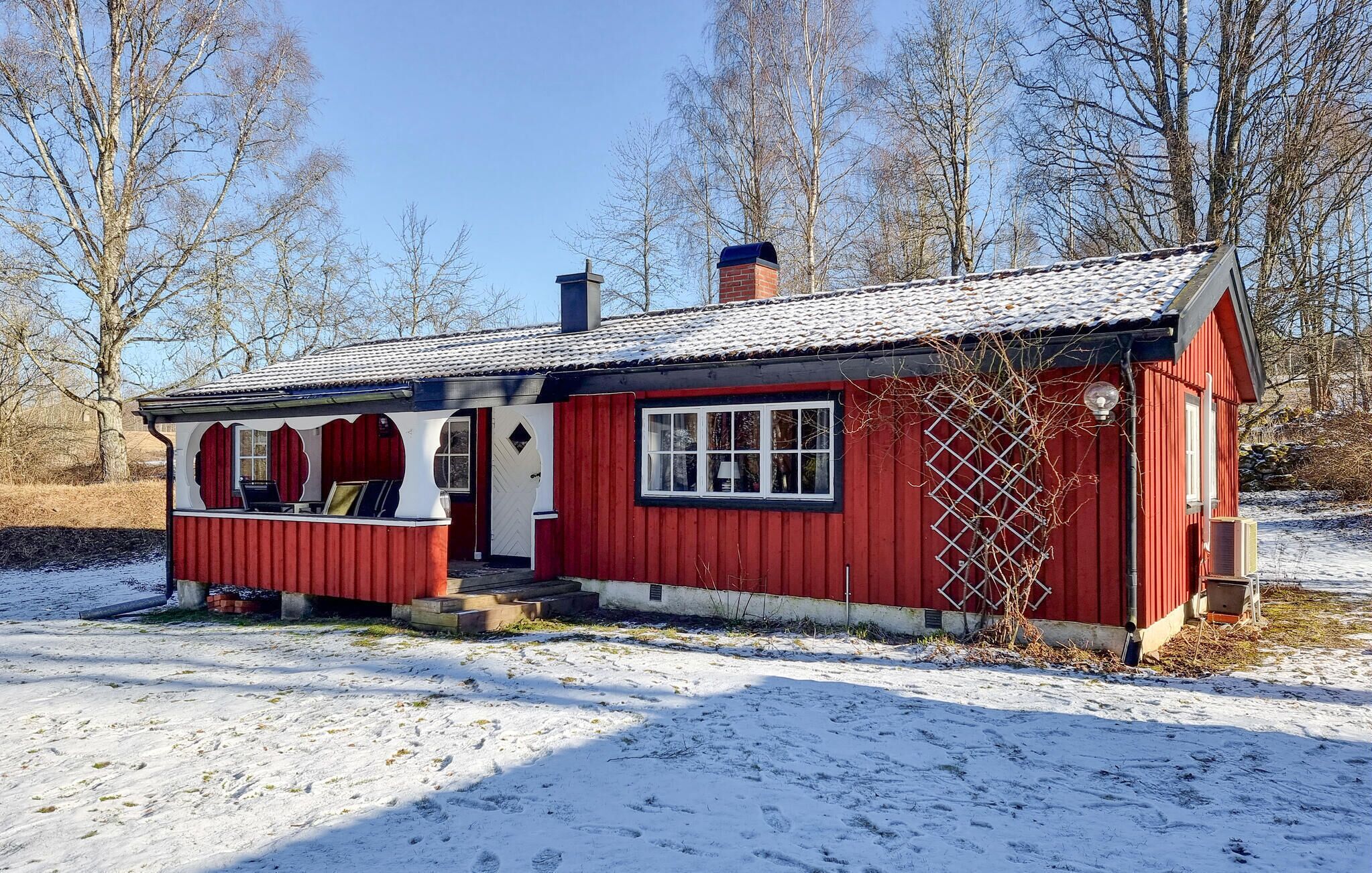 A small, red wooden cabin with a single window and a chimney, surrounded by snow and trees.