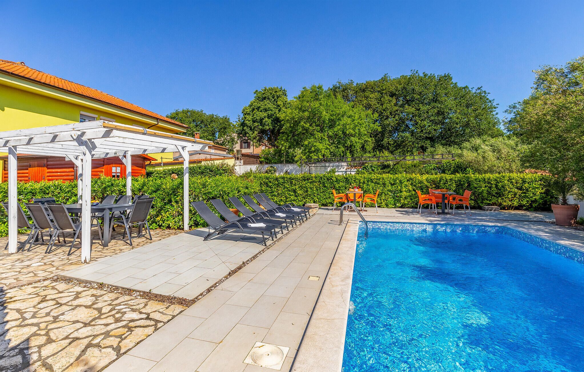 A swimming pool with lounge chairs and tables, surrounded by greenery and a building with a yellow facade.