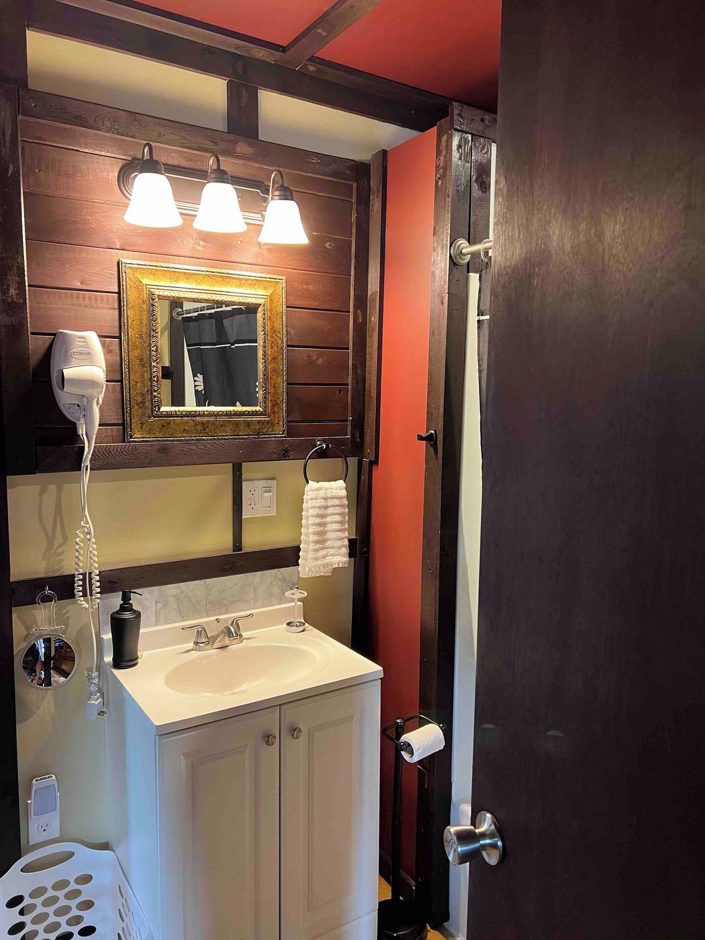 Ein Badezimmer mit einem Holzkosmetikschrank, einem Spiegel mit goldener Rahmung, einem Fön, einem Handtuchhalter, einem Waschbecken und einem Toilettenpapierhalter.