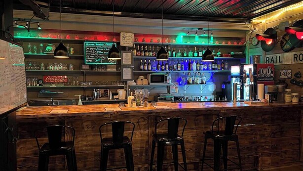 Een bar met een houten toonbank, zwarte barstoelen en planken vol flessen en glazen.