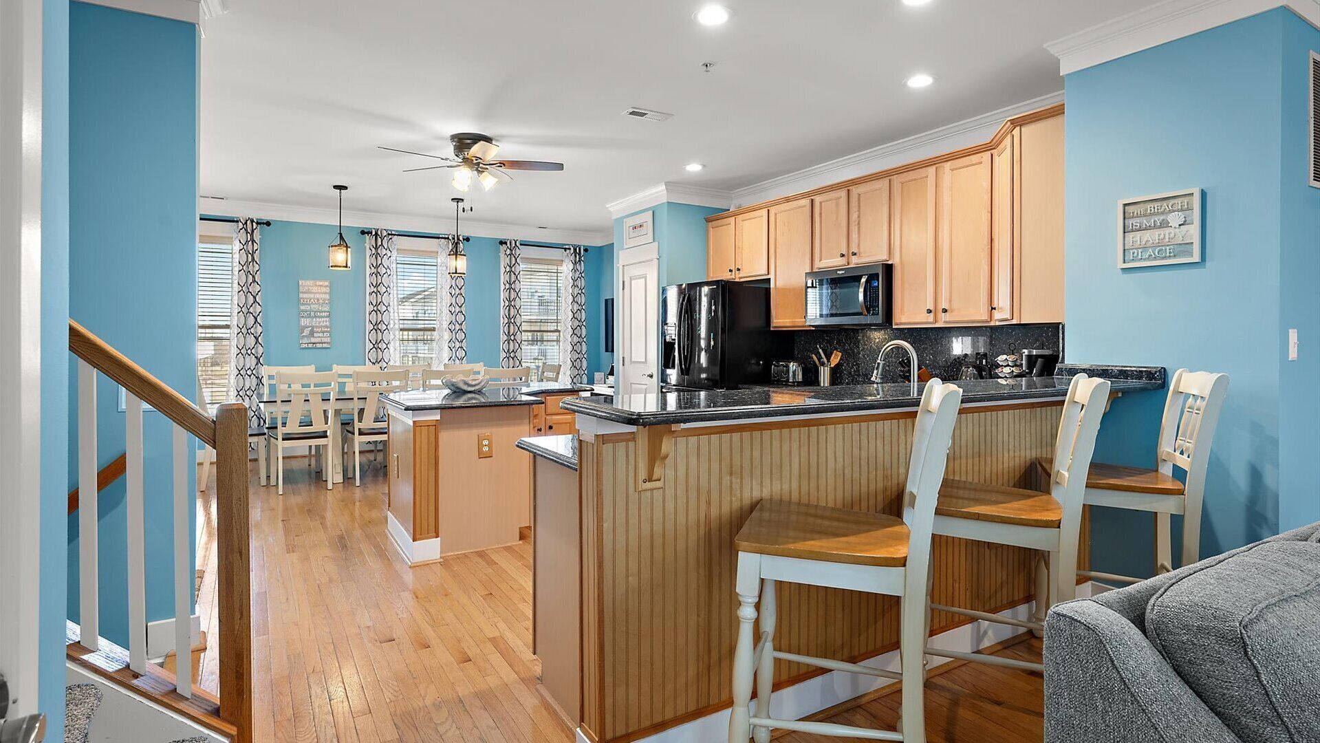 A modern kitchen with wooden cabinets, a black refrigerator, and a microwave. There is a dining area with white chairs and a wooden table.