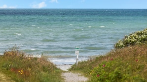 En kyststi der fører ned til havet, med et skilt der viser et telefonnummer og et symbol for en redningsstation.