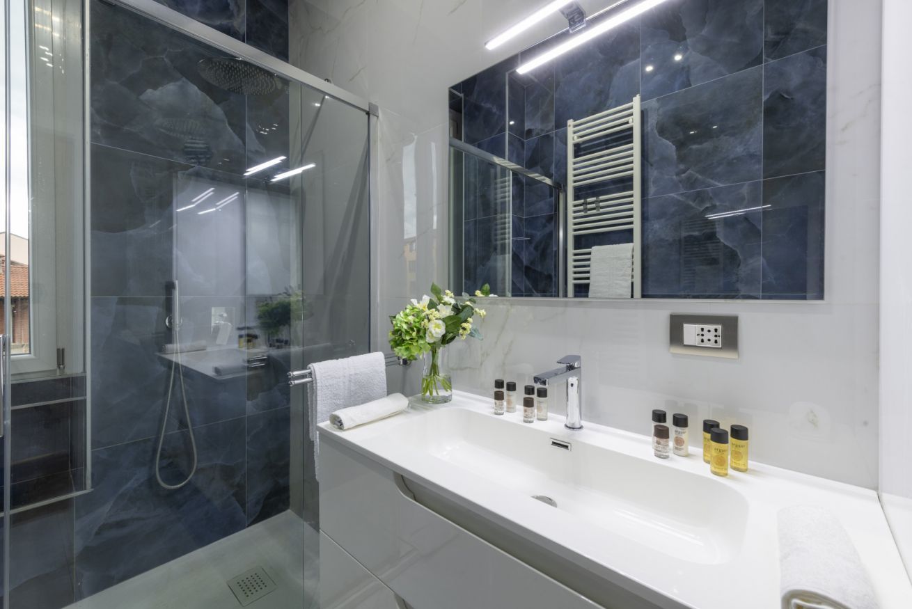 Een moderne badkamer met een douchecabine van glas, een wit wastafelmeubel met toiletartikelen en een marmeren spatwand.