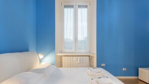 A bedroom with a bed, a window with curtains, a radiator, and a bedside table with a lamp. - GIACOSA, 71 – Spacious apartment for 6 (Milano)