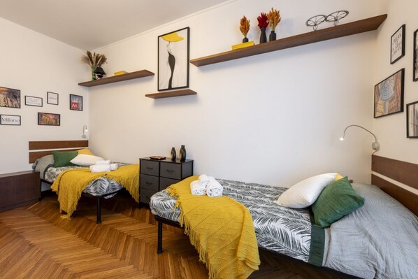 Una camera con due letti, pavimento in legno, un comodino, una cassettiera e mensole a parete con opere d'arte e oggetti decorativi.