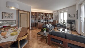 A modern living room with a sofa, coffee table, and dining area with a table set for breakfast. There is a bookshelf filled with books and a TV in the corner. - Otto Novembre, 3 – Spacious and Elegant with a View (Milano)